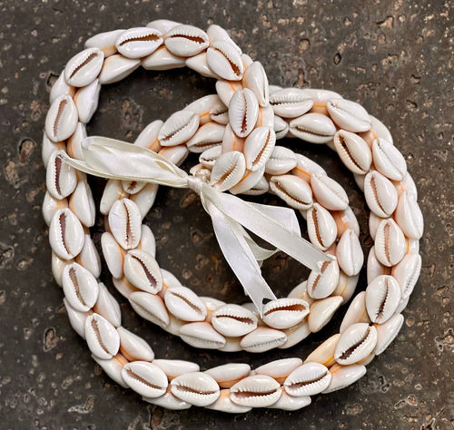 Braided cowry shell lei with a white ribbon on a textured surface