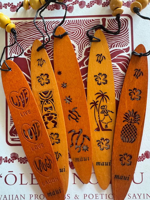 A collection of wooden bookmarks with Hawaiian inspired designs, featuring phrases and images related to Hawaii, displayed on a surface with a floral patterned book in the background.