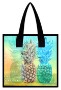 Reusable tote bag featuring two pineapples with a tropical watercolor-style background in orange, green, and blue hues. The bag design includes words like 'Aloha,' 'Hawaii,' 'Waikiki,' and names of Hawaiian islands subtly overlaid. Black handles and trim complete the design.