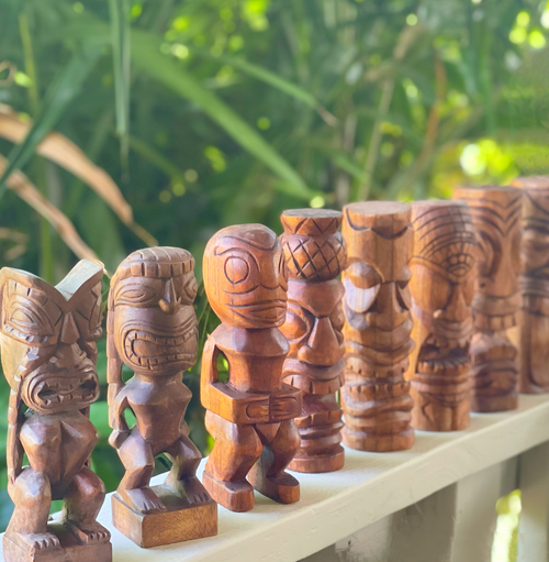 A collection of hand-carved wooden tikis displayed in a row, each with unique facial features and designs, set against a blurred background of lush greenery.