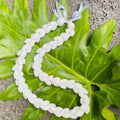 A braided bubble shell lei with kukui nuts, featuring white bubble shells and a white satin ribbon tie, displayed on a green leafy background.