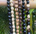A selection of kukui nut lei necklaces in various colors displayed on a wooden rack, with greenery in the background.